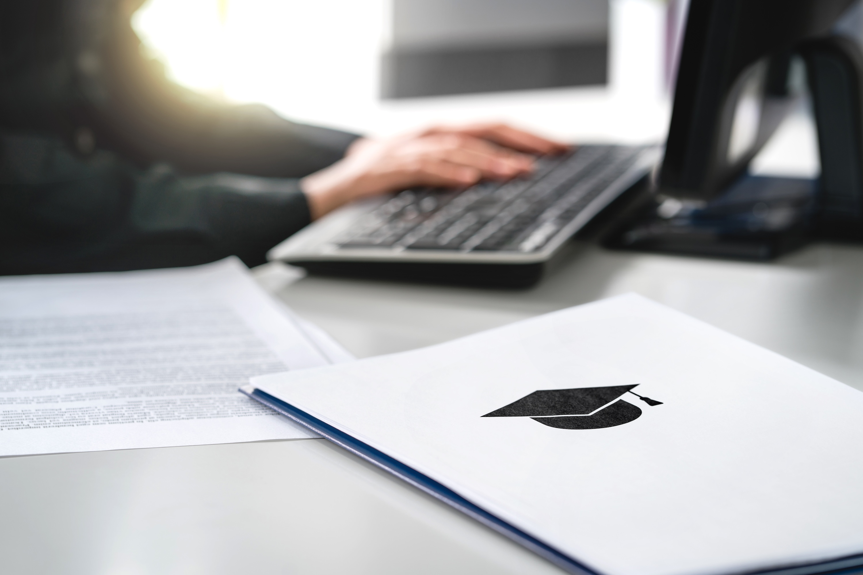 Hände auf Tastatur, im Bildvordergrund Unterlafen mit Doktorhut-Symbol